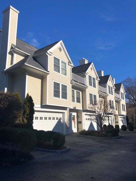 A row of light yellow townhouses with dark shingled roofs and white garage doors under a clear blue sky.