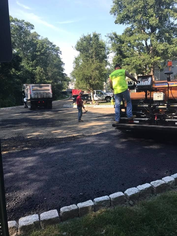 Workers use an asphalt paver to lay fresh blacktop on a driveway lined with stone pavers.