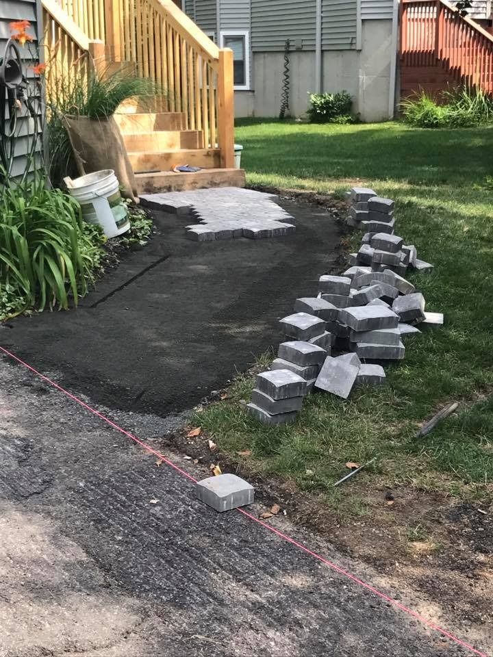 A partially paved walkway under construction next to a wooden staircase, with piles of grey pavers on the adjacent grass.