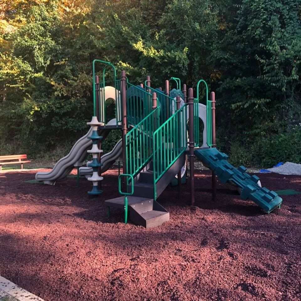 A green and tan playground structure with stairs, two slides, and railings, on a dark wood-chip surface.