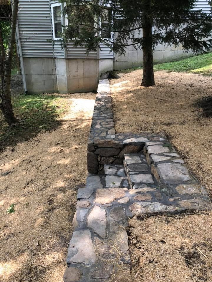 A stone retaining wall with integrated steps built into a sloped, wood-chipped yard next to a house.