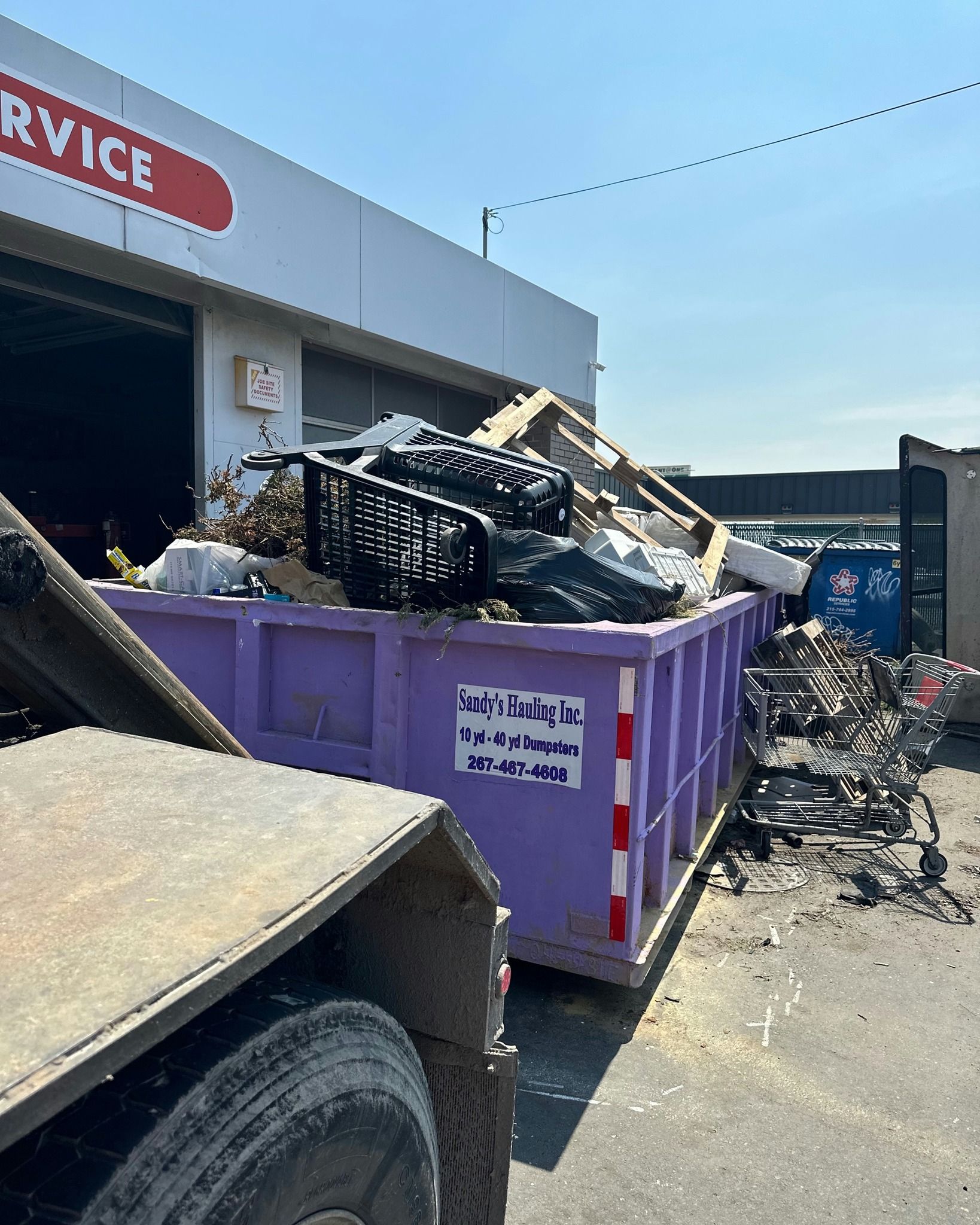 Purple dumpster filled with debris next to a building; truck in foreground.