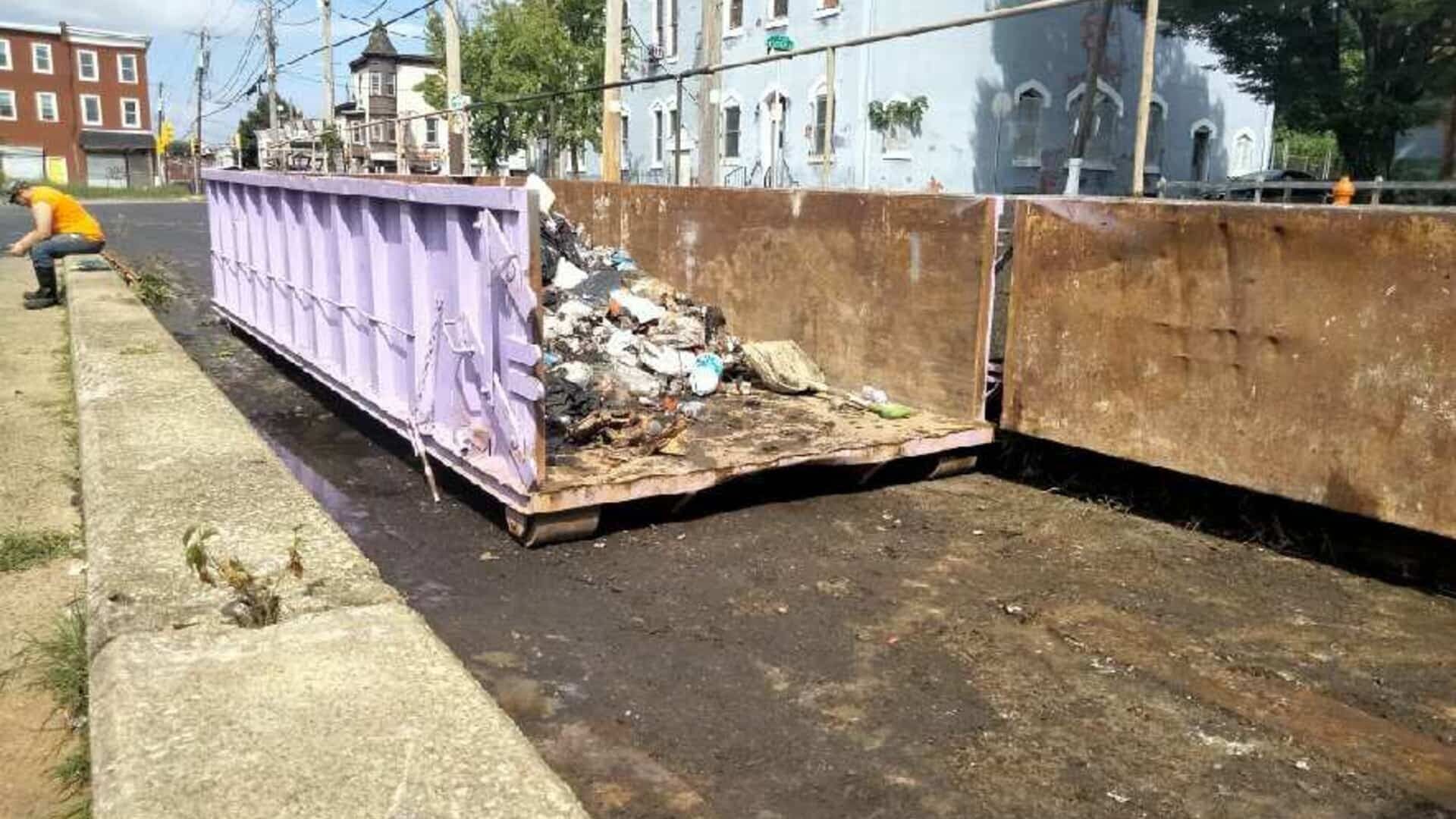 Purple and rust-colored dumpsters overflowing with trash on a street, a person in orange shirt nearby.
