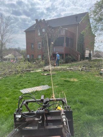 Lawn tractor in foreground facing a brick house, with a person near the porch and storm debris in the yard.