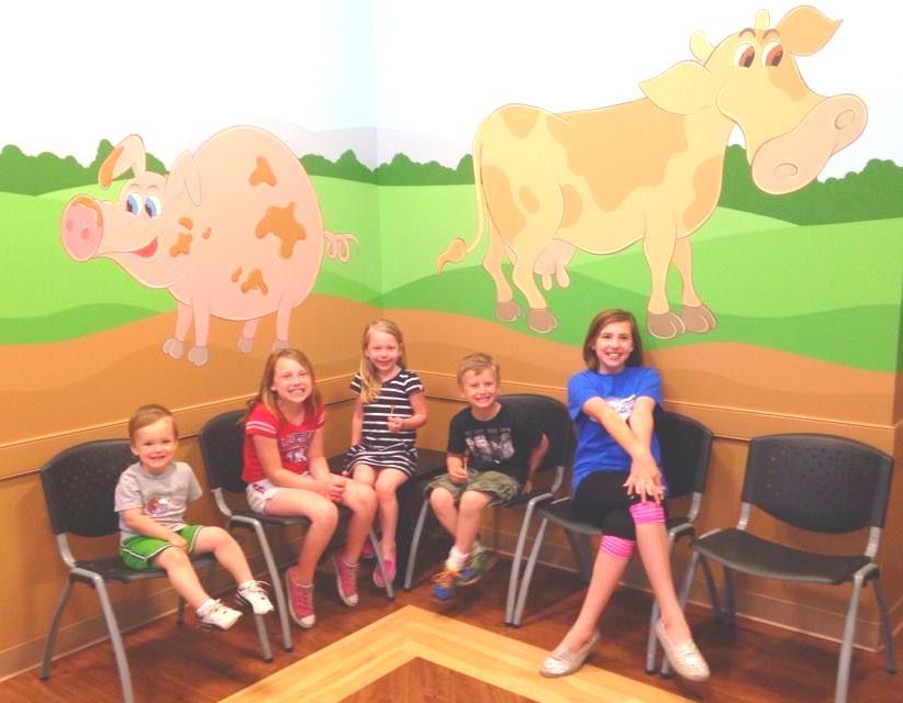 A group of children are sitting in a waiting room with a drawing of a cow and pig on the wall.