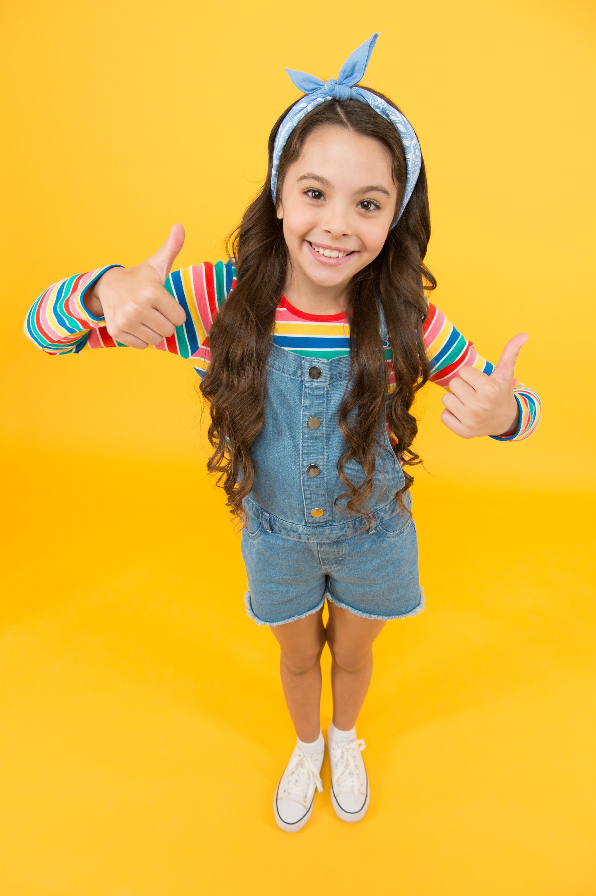 A girl in a colorful shirt with yellow background is giving two thumbs up.