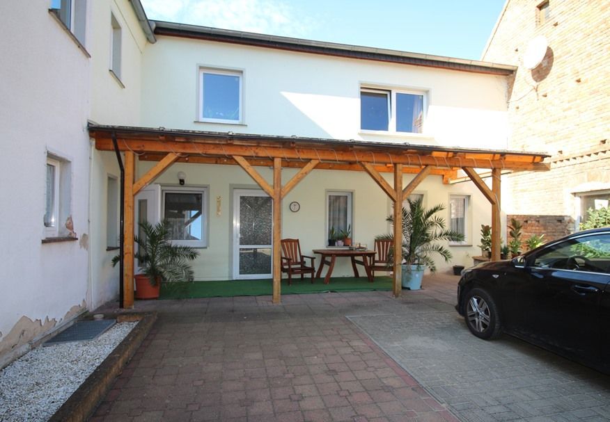 Innenhof mit hölzernem Carport, Terrassenmöbeln, Topfpflanzen und einem schwarzen Auto neben einem weißen Haus