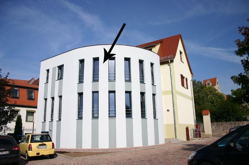 Weißes, geschwungenes Gebäude mit hohen, schmalen Fenstern und rotem Dach; ein Pfeil zeigt auf ein oberes Fenster.