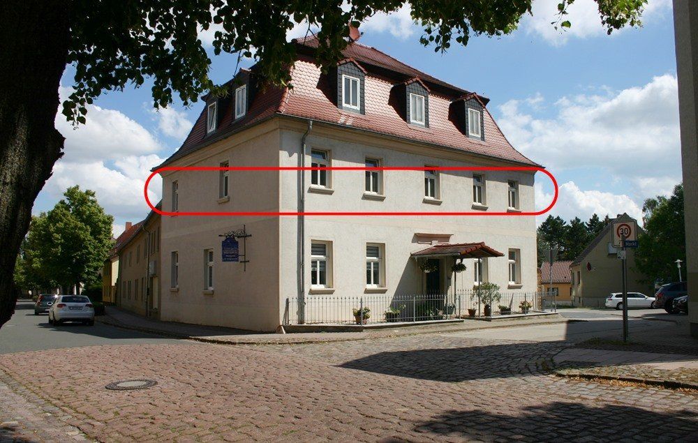 Zweistöckiges cremefarbenes Gebäude mit rotem Dach und rot umrandeten oberen Fenstern an einer Kopfsteinpflasterstraße