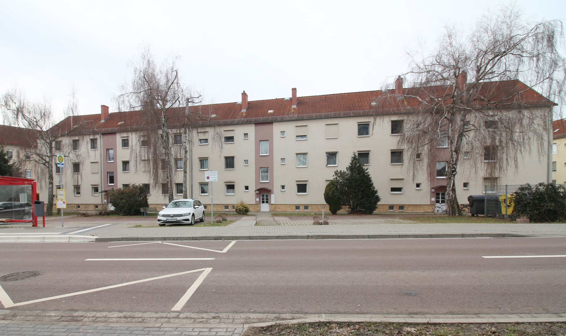 Mehrfamilienhaus mit rotem Dach hinter einer breiten Straße und geparkten Autos an einem bewölkten Tag
