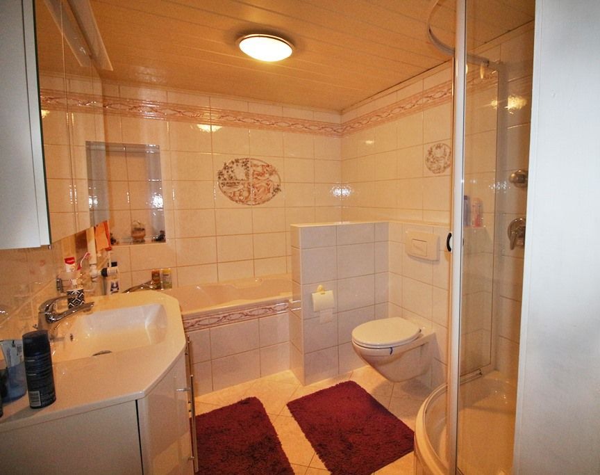 Kleines Badezimmer mit Waschbecken, Toilette, Glasdusche und roten Teppichen unter warmem Licht