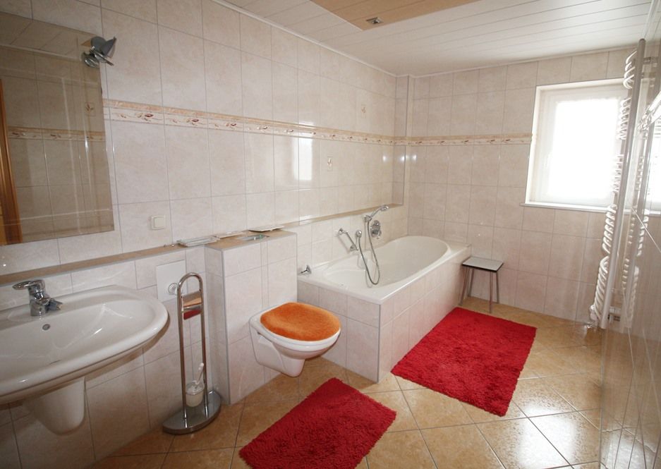 Helles, gefliestes Badezimmer mit Badewanne, Toilette, Waschbecken, Spiegel und roten Badematten.