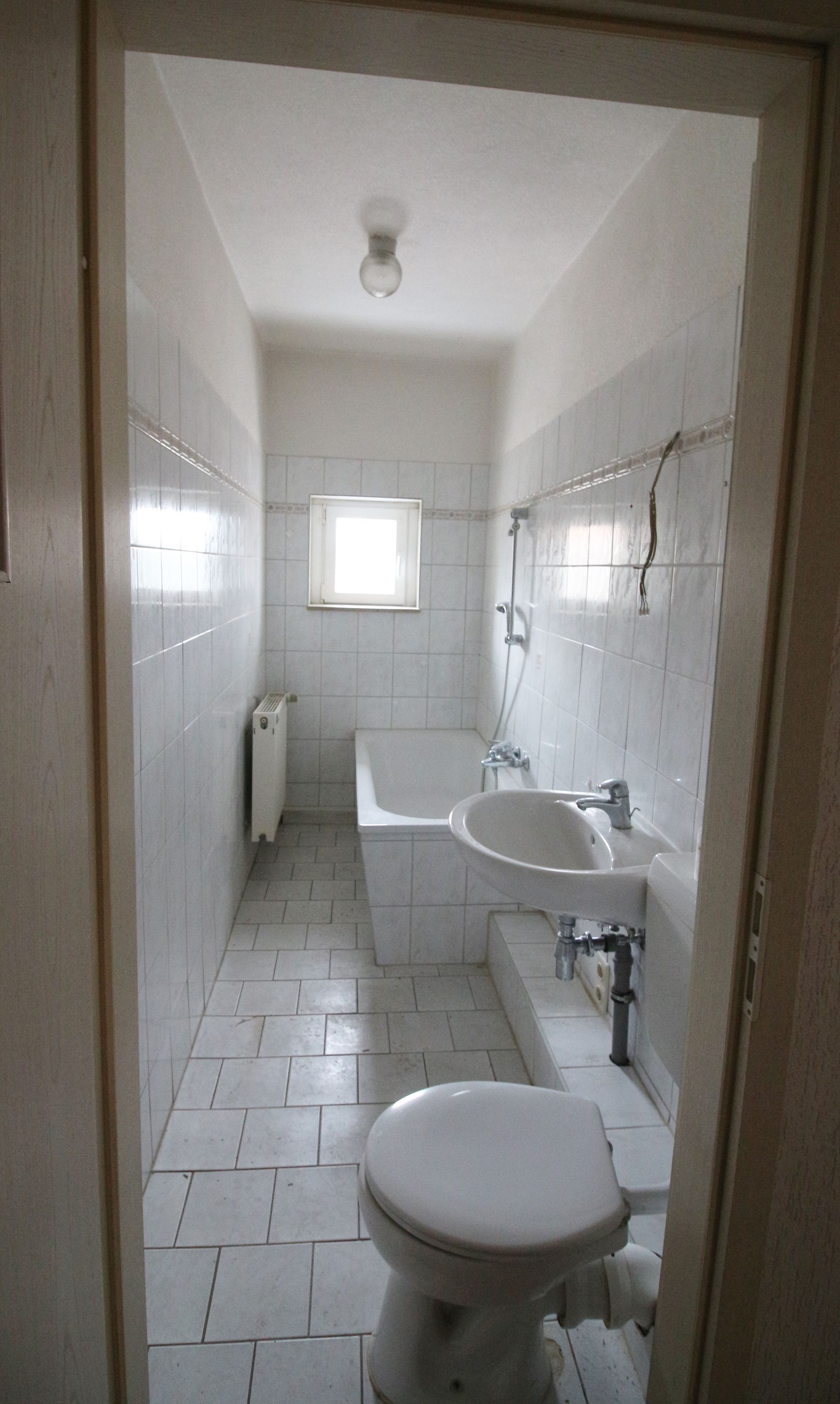 Schmale, weiße Badezimmer mit Waschbecken, Toilette, gefliesten Wänden und einem kleinen Fenster am Ende.