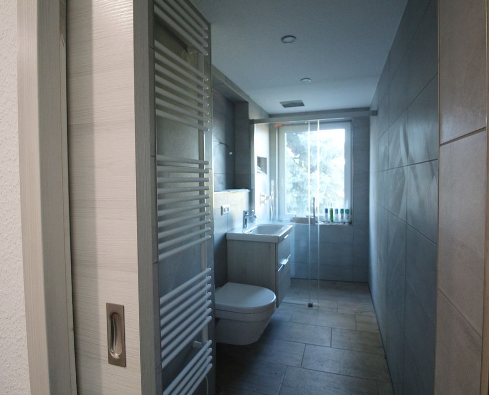 Schmaler Badezimmerflur mit Toilette, Waschbecken, gefliesten Wänden und einem Fenster am anderen Ende
