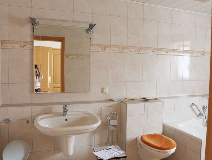 Helles, gefliestes Badezimmer mit Waschbecken, Spiegel, Toilette und Badewanne mit orangefarbenem Sitzbezug