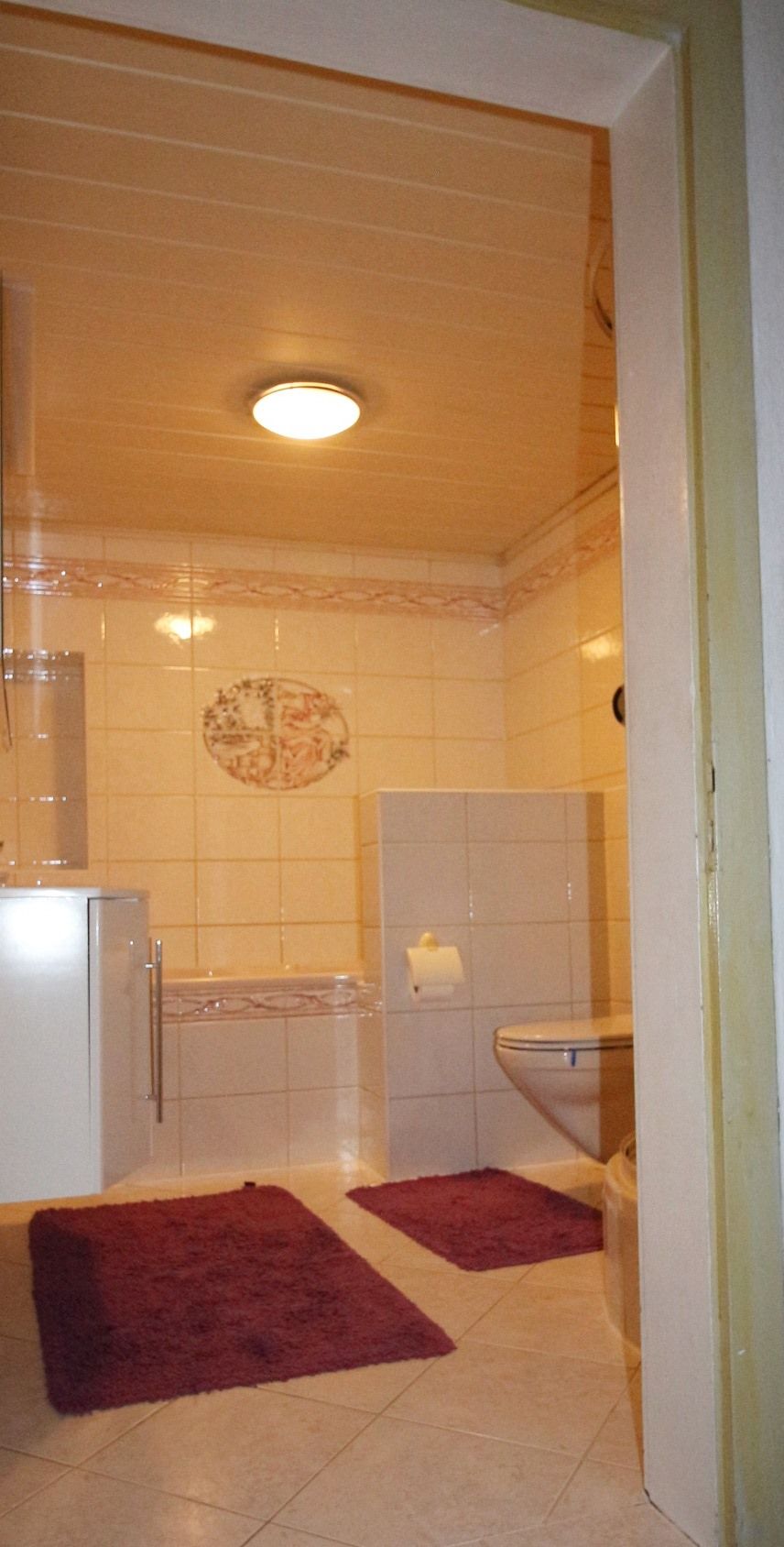 Kleines Badezimmer mit beigen Wänden, weißen Fliesen, Waschbecken und zwei dunkelvioletten Badematten.