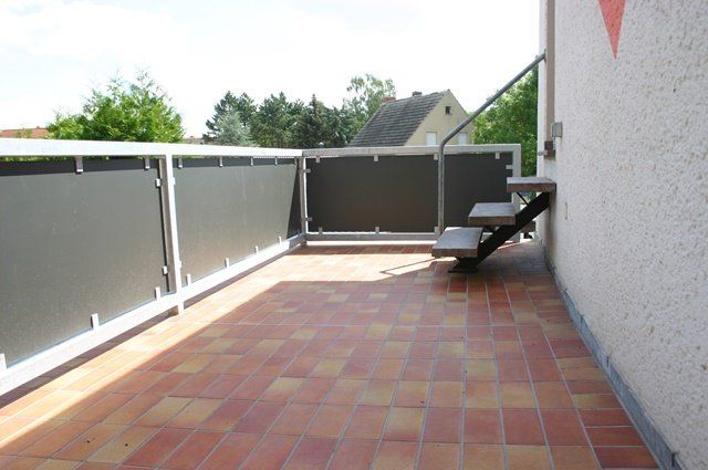 Ein langer, gefliester Balkon mit grauem Geländer, weißer Wand und einem schwarzen Stuhl am hinteren Ende.