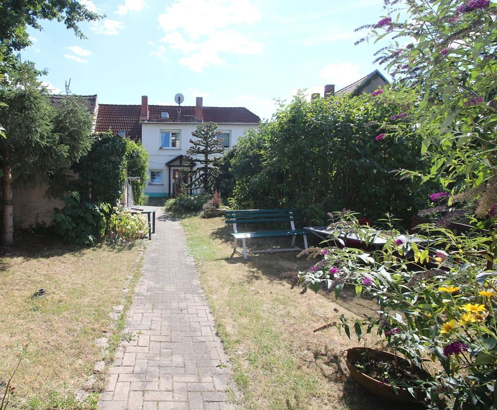 Ein Gartenweg führt zu einem weißen Haus, zu dessen beiden Seiten Bänke und blühende Sträucher stehen.