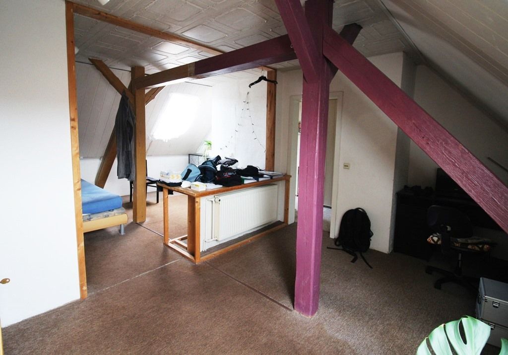 Kleines Dachzimmer mit freiliegenden Balken, Schreibtisch, Heizkörper und schrägen weißen Wänden unter einer niedrigen Decke