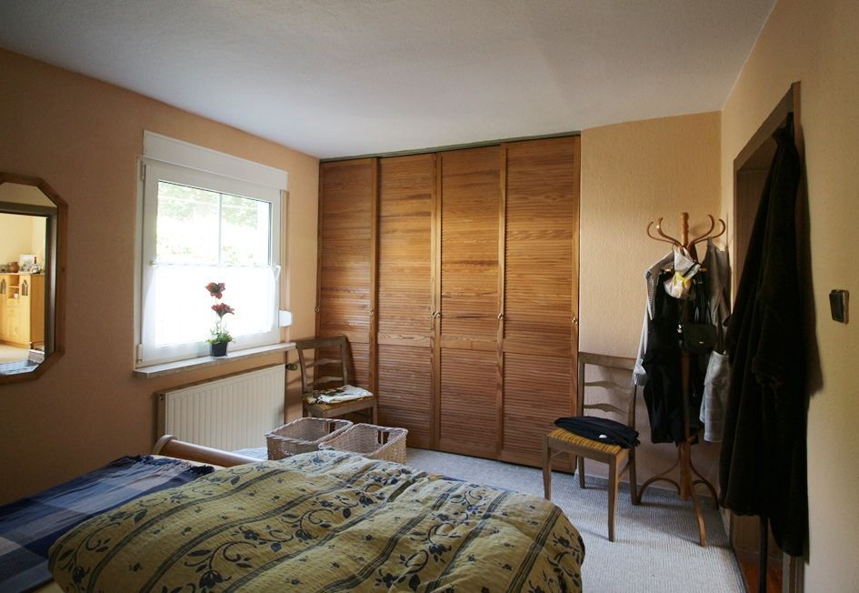 Kleines Schlafzimmer mit Bett, Fenster, Holzschrank, Stuhl und Garderobenständer.