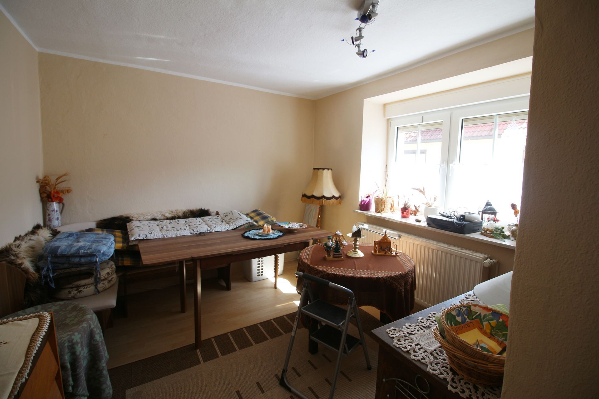 Kleines Esszimmer mit Tisch, Stühlen, Bett, Lampe und Fenster mit Heizkörper darunter.