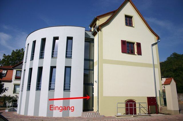 Modernes weiß-cremefarbenes Gebäude mit roter Tür und Fenstern; roter Pfeil zeigt auf den Eingang (