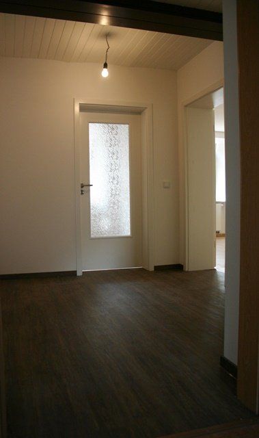 Leerer Raum mit dunklem Holzboden, weißen Wänden und einer Milchglastür unter einer hängenden Glühbirne.