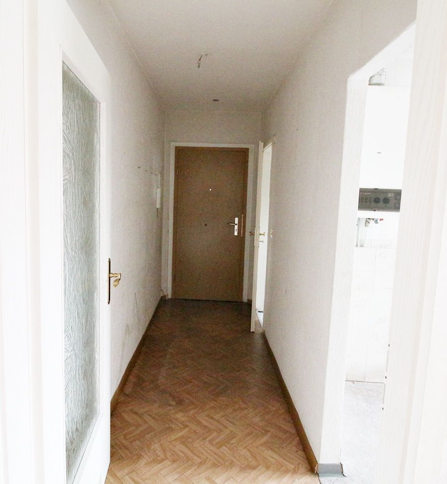 Schmaler Flur mit weißen Wänden, Holzboden und einer geschlossenen braunen Tür am Ende.