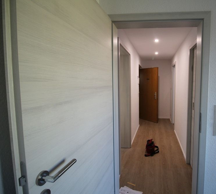 Eine offene weiße Tür gibt den Blick auf einen schmalen Flur mit Holzboden frei, auf dem Boden steht eine Tasche.
