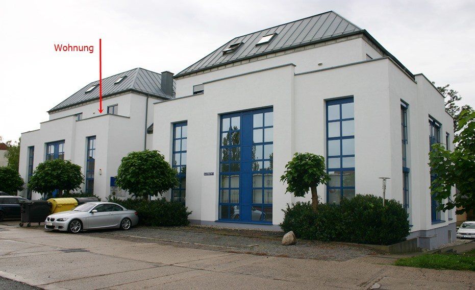 Modernes weißes Geschäftsgebäude mit blauen Türen und Fenstern, geparkten Autos und einem roten Schild draußen.