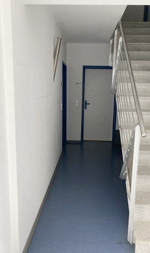 Schmaler Flur mit blauem Boden, weißen Wänden, Treppengeländer auf der rechten Seite und einer geschlossenen Tür geradeaus.