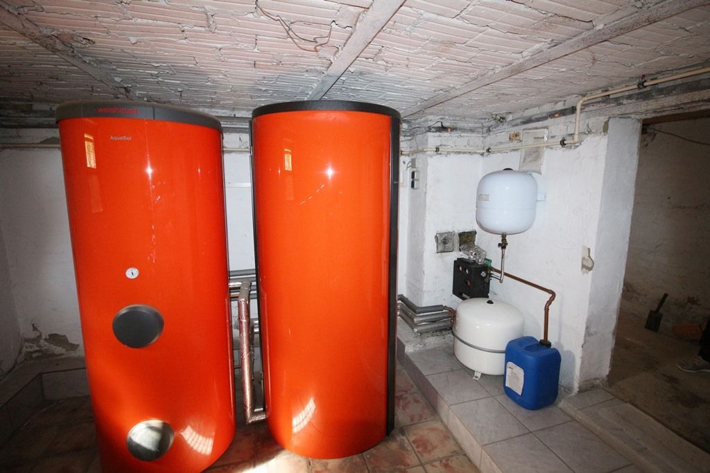 Heizraum mit zwei hohen orangefarbenen Wassertanks, Rohren und einer blauen Pumpe auf Betonboden.
