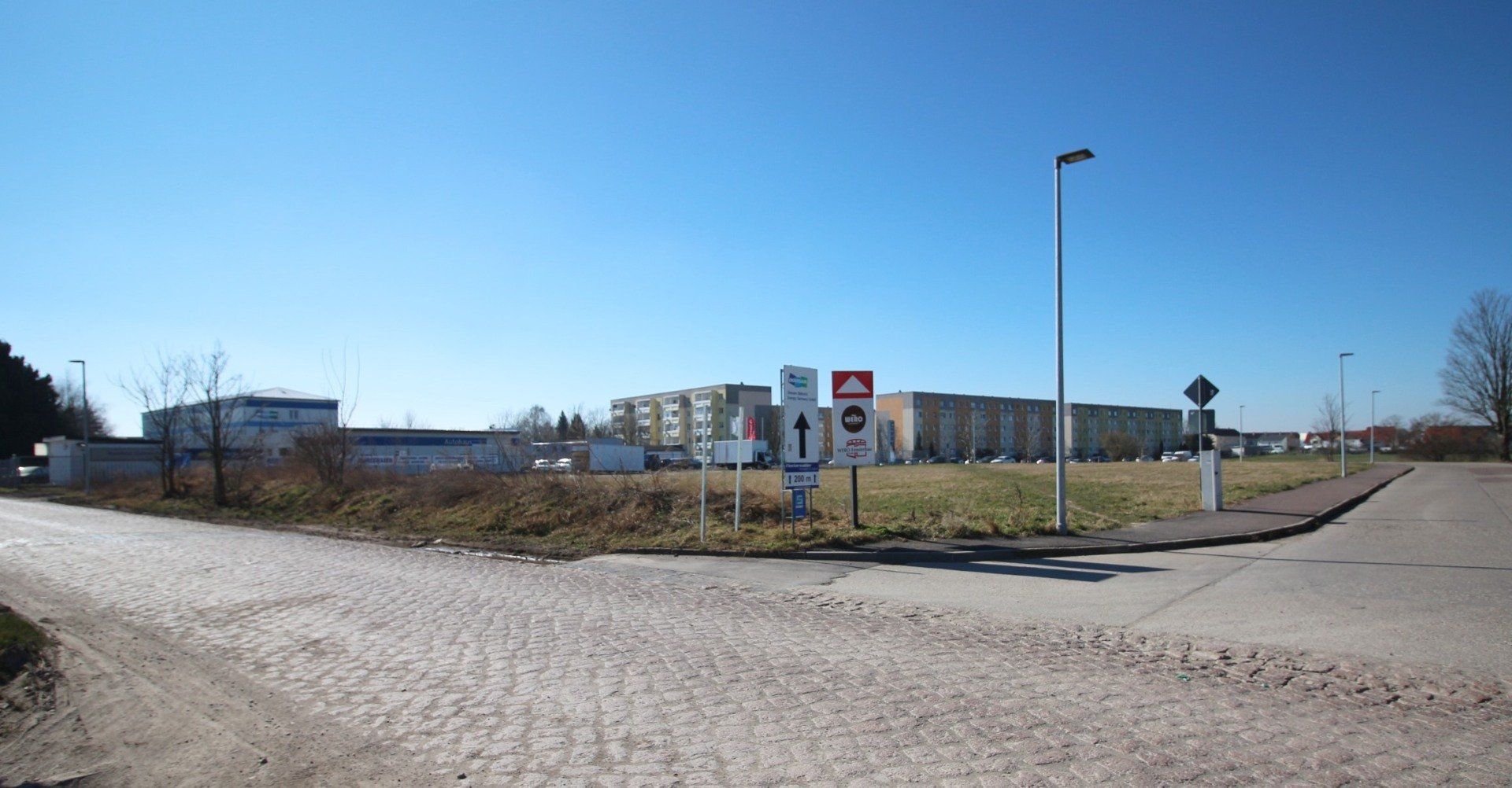 Kreuzung einer Schotterstraße mit Verkehrsschildern und Straßenlaternen neben niedrigen Gebäuden unter klarem, blauem Himmel