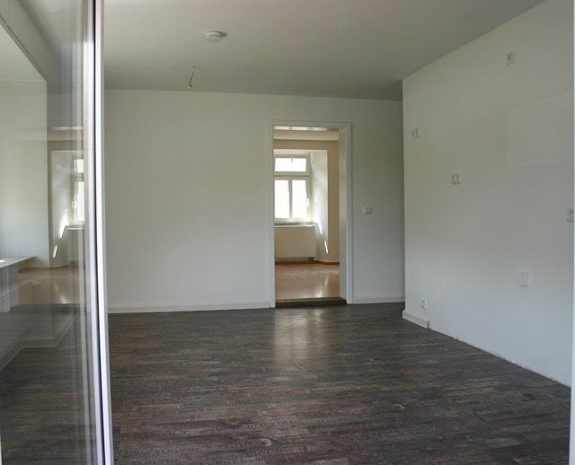 Ein leerer, weißer Raum mit dunklem Holzboden und einer Tür zu einem helleren angrenzenden Raum.