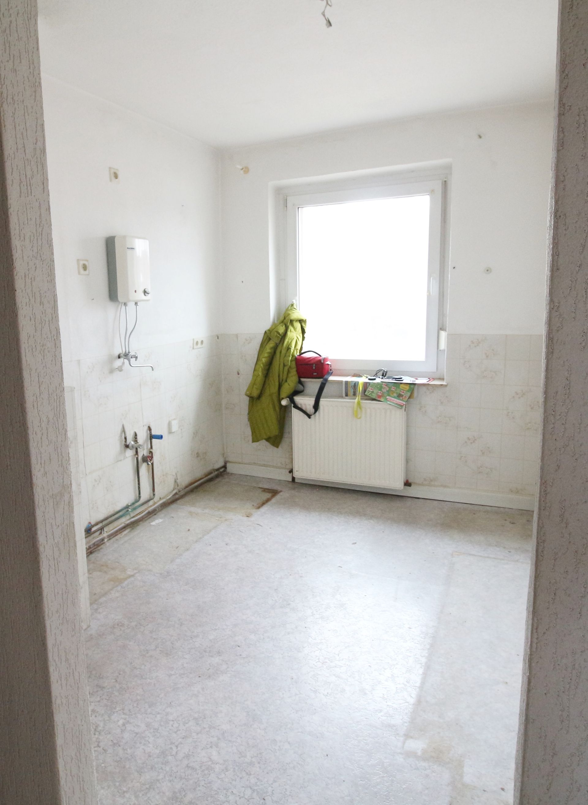 Leere kleine Küche mit weißen Wänden, einem Fenster, einem Spülbecken und einer grünen Jacke, die am Heizkörper hängt.