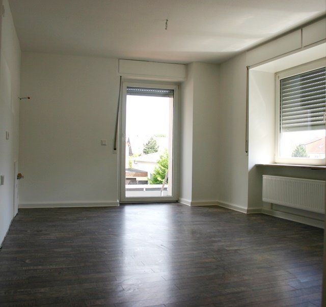Leeres Zimmer mit dunklem Holzboden, weißen Wänden, offener Tür und Fenster mit Jalousien.