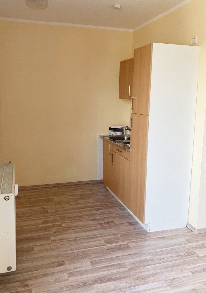 Kleiner, unmöblierter Raum mit Holzboden und beigen Wänden, rechts davon eine Küchenzeile und ein Schrank.