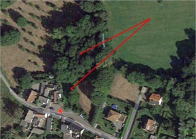Luftbild einer Rasenfläche mit Häusern, rote Linien markieren einen Pfad durch Bäume und offenes Feld.