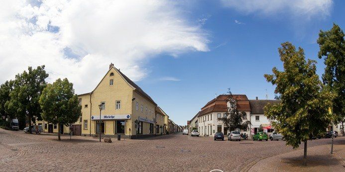 Ein breiter, belegter Platz mit gelben und weißen Gebäuden, Bäumen und einigen Menschen unter blauem Himmel