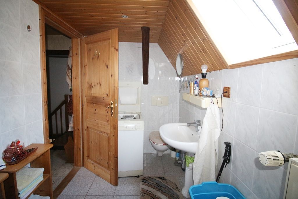 Kleines Dachgeschossbad mit schräger Holzdecke, offener Holztür, Waschbecken, Toilette und gefliesten Wänden.