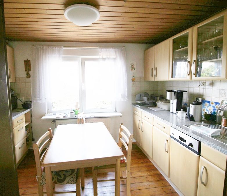 Helle Küche mit Holzdecke, weißen Schränken und einem Esstisch an einem sonnendurchfluteten Fenster.