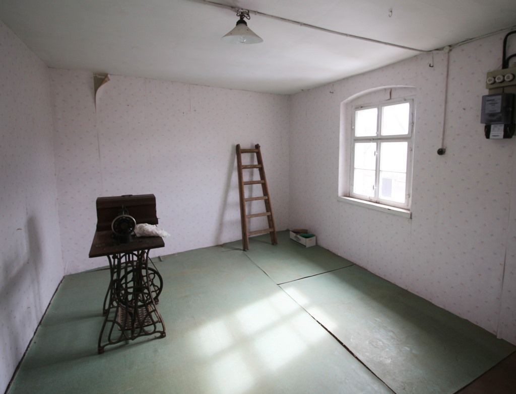 Leeres Zimmer mit grünen Fußmatten, Leiter, kleinem Ofen und Sonnenlicht durch ein Fenster
