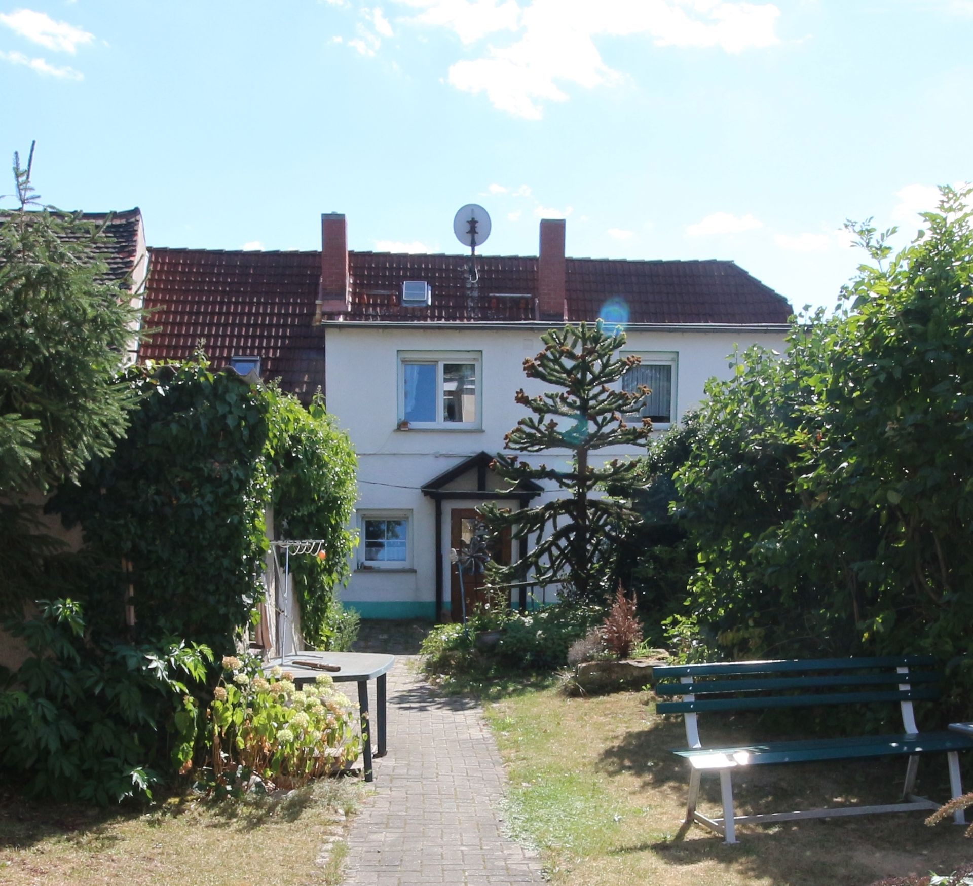 Kleines Haus mit rotem Dach und Gartenweg, Ansicht von vorne, im Vordergrund eine Bank.