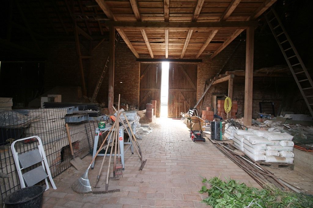 Düsteres Scheuneninterieur mit Werkzeugen, gestapelten Materialien und einer hellen, offenen Tür am anderen Ende