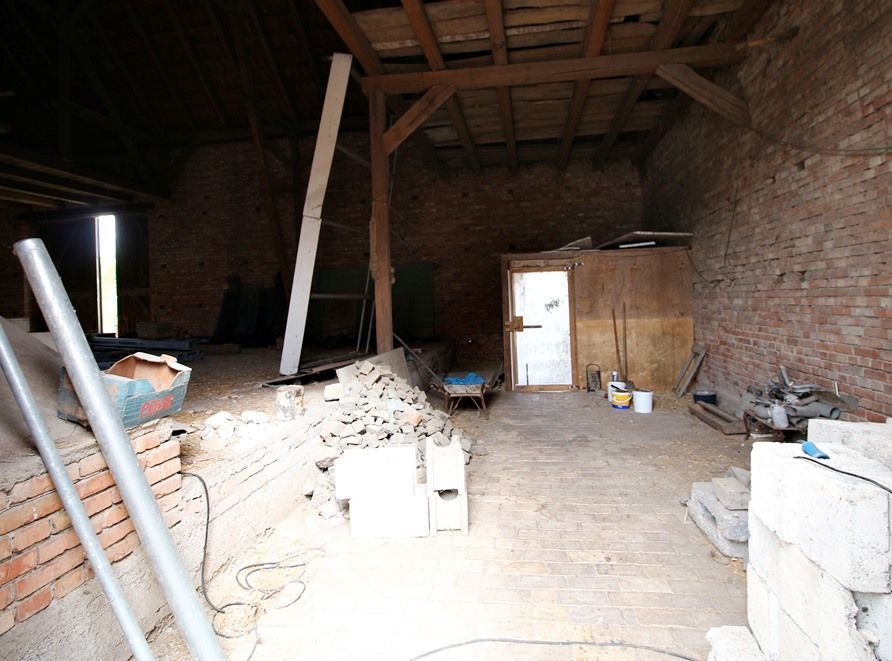 Unfertiger Dachboden mit freiliegendem Mauerwerk, Holzbalken, Bauschutt und einem hellen Türdurchgang an der Rückseite