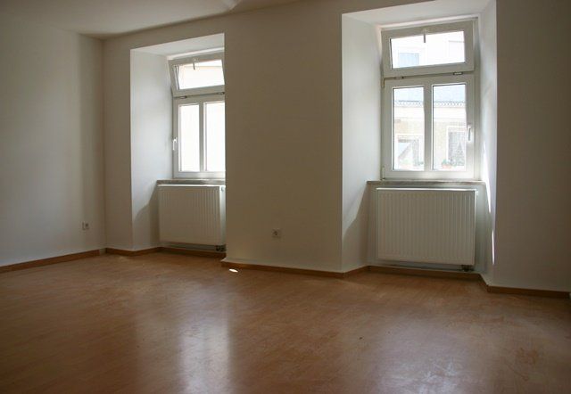 Leerer Raum mit Holzboden, weißen Wänden und zwei hohen Fenstern mit Heizkörpern darunter.