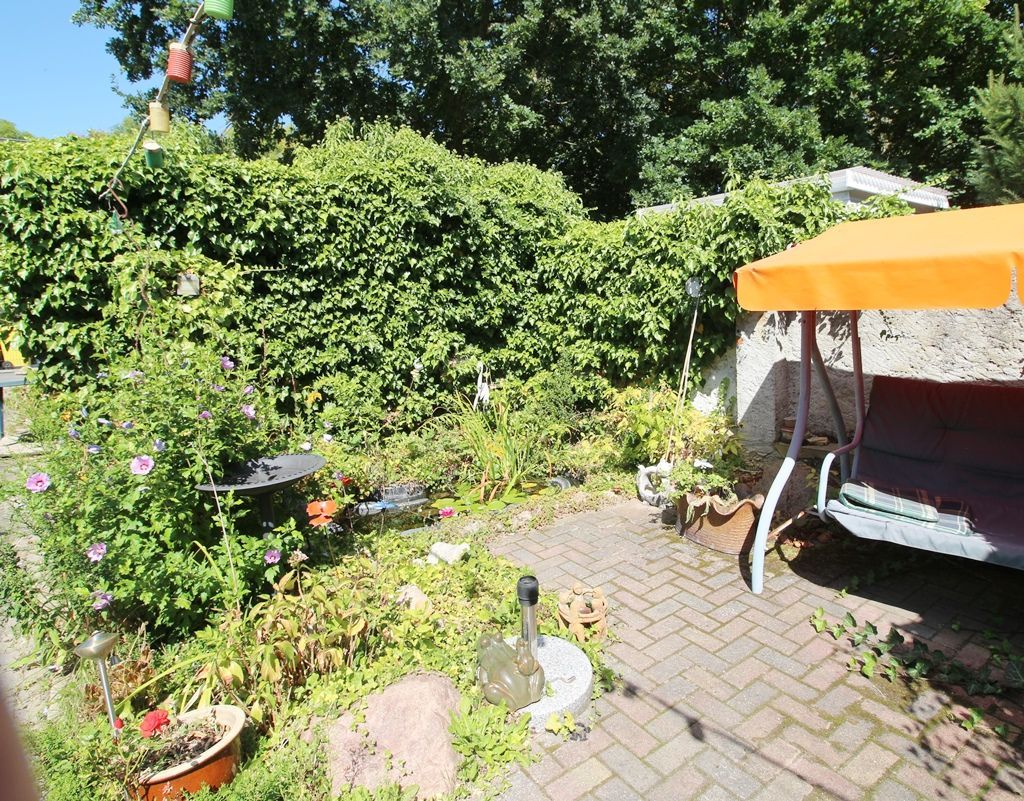 Sonnige Gartenterrasse mit grünen Hecken, Ziegelpflaster, Topfblumen und einer orangefarbenen Schaukelbank mit Sonnendach