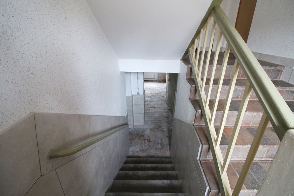 Treppenhaus mit abgenutzten Stufen, beigen Wänden und einem Metallgeländer, das zu einer Tür hinunterführt.