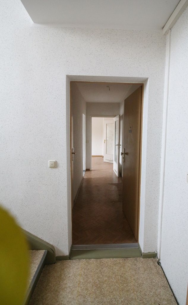 Schmaler Flur mit weißen Wänden, braunem Boden und einer Tür, die in einen helleren Raum führt.