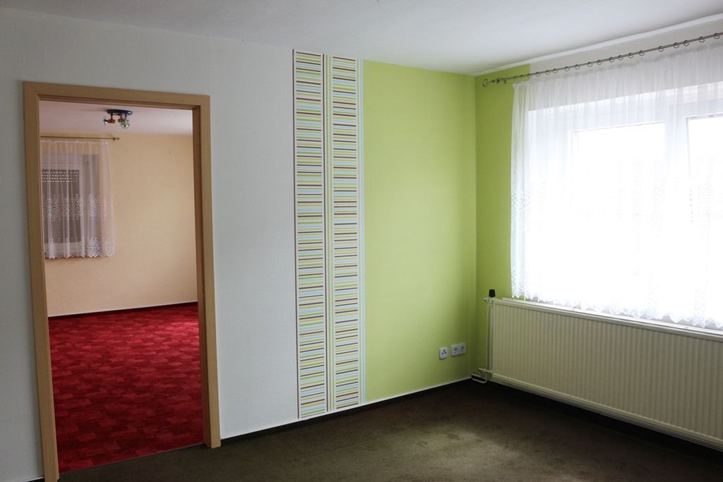 Ein leerer Raum mit grün-weißen Wänden, dunklem Teppich und einer Tür zu einem rot ausgelegten Raum.
